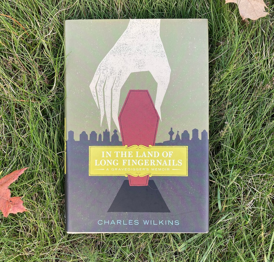 A book lying in the grass. The books title is "In the Land of Long Fingernails: A Gravedigger's Memoir" by Charles Wilkins.